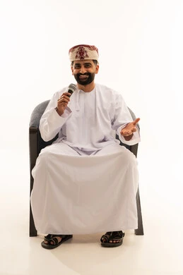 The art of delivering speeches and seminars, vocal training, and presenting radio programs, a portrait of an Arab Gulf Omani man wearing a dishdasha and a turban sitting on a chair holding a microphone in his hands, gestures and facial expressions conveying singing and music, white background.