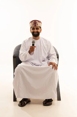 التدريب الصوتي وتقديم البرامج الاذاعيه، فن إلقاء الخطابات والندوات، بورتريه لرجل عربي خليجي عماني يرتدي الدشداشة و العمامة يجلس على الكرسى يمسك بيدية الميكروفون، ايماءات وتعابير وجه و يدين تعبر عن الغناء والطرب، خلفية بيضاء