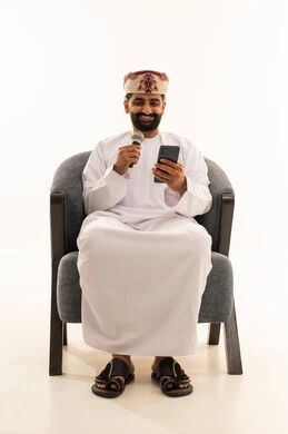 تقديم العرض المسرحي، ممارسة هواية الغناء للتسلية والترفيه، مواهب فنية في عالم الموسيقى، الفن المسرحي الغنائي، ببورتريه لرجل عربي خليجي عماني يرتدي الدشداشة و العمامة يجلس على الكرسى يحمل بيده الميكروفون و الهاتف المحمول ، خلفية بيضاء