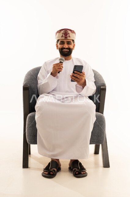ممارسة هواية الغناء للتسلية والترفيه، مواهب فنية في عالم الموسيقى، الفن المسرحي الغنائي، تقديم العرض المسرحي، ببورتريه لرجل عربي خليجي عماني يرتدي الدشداشة و العمامة يجلس على الكرسى يحمل بيده الميكروفون و الهاتف المحمول ، خلفية بيضاء