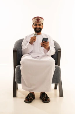 Practicing the hobby of singing for fun and entertainment, artistic talents in the world of music, musical theater art, presenting the theatrical performance, with a portrait of an Arab Gulf Omani man wearing a dishdasha and a turban sitting on a chair holding a microphone and a mobile phone, white background.
