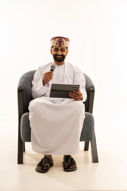 الاندماج مع الأغنية، تسجيل مقطع صوتي غنائي، مواهب غنائية فنية، بورتريه لرجل عربي خليجي عماني يرتدي الدشداشة و العمامة يجلس على الكرسى يقوم بالغناء باستخدام الميكروفون يمسك بيدية التابلت بتعابير الحماس، خلفية بيضاء