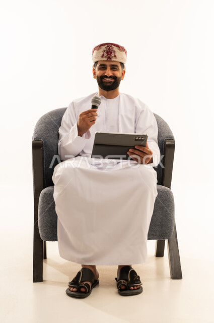 تسجيل مقطع صوتي غنائي، مواهب غنائية فنية، الاندماج مع الأغنية، بورتريه لرجل عربي خليجي عماني يرتدي الدشداشة و العمامة يجلس على الكرسى يقوم بالغناء باستخدام الميكروفون يمسك بيدية التابلت بتعابير الحماس، خلفية بيضاء