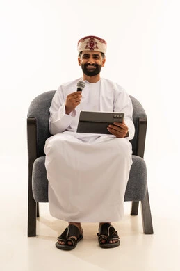 تسجيل مقطع صوتي غنائي، مواهب غنائية فنية، الاندماج مع الأغنية، بورتريه لرجل عربي خليجي عماني يرتدي الدشداشة و العمامة يجلس على الكرسى يقوم بالغناء باستخدام الميكروفون يمسك بيدية التابلت بتعابير الحماس، خلفية بيضاء