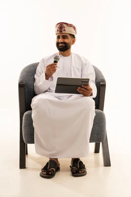 مواهب غنائية فنية، تسجيل مقطع صوتي غنائي، الاندماج مع الأغنية، بورتريه لرجل عربي خليجي عماني يرتدي الدشداشة و العمامة يجلس على الكرسى يقوم بالغناء باستخدام الميكروفون يمسك بيدية التابلت بتعابير الحماس، خلفية بيضاء