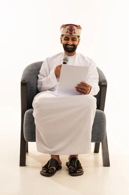 اعداد التقارير الخاصة بالعمل، تقديم الفقرات باستخدام الميكروفون، متابعة ومراجعة الخطط والمشاريع المكتبية، بورتريه لرجل عربي خليجي عماني يرتدي الدشداشة و العمامة يجلس على الكرسى ينظر الى الاوراق البيضاء يمسك بيده المايك, التدقيق في قراءة الملاحظات، خلفية بيضاء