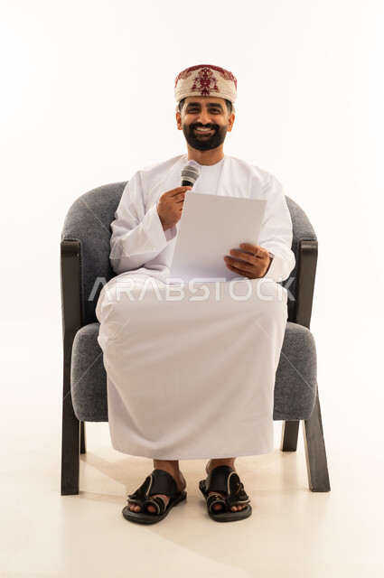 النظر للكاميرا بتعابير السرور، اعداد التقارير الخاصة بالعمل، تقديم الفقرات باستخدام الميكروفون، متابعة ومراجعة الخطط والمشاريع المكتبية، بورتريه لرجل عربي خليجي عماني يرتدي الدشداشة و العمامة يجلس على الكرسى ينظر الى الاوراق البيضاء يمسك بيده المايك, التدقيق في قراءة الملاحظات، خلفية بيضاء
