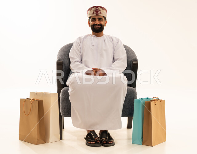 Taking advantage of discounts and clearance sales, shopping for necessary and essential items, discount seasons in the Sultanate of Oman, a portrait of an Arab Gulf Omani man wearing a dishdasha and a turban sitting on a chair with paper bags beside him, looking at the camera with a joyful expression, white background.