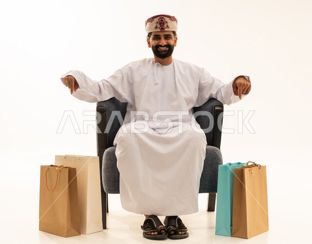 حركات تدل على هذا المكان (هنا)، التبضع وأخذ الحاجات اللازمة والضرورية, مواسم التخفيضات في سلطنة عمان , الاستفادة من الحسومات والتصفيات, بورتريه لرجل عربي خليجي عماني يرتدي الدشداشة و العمامة يجلس على الكرسى و بجانبه أكياس ورقية يشير بإصبعه نحو الأسفل , خلفية بيضاء