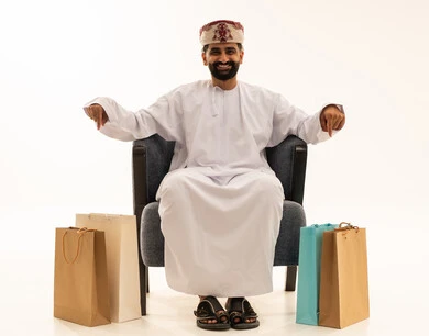 Movements indicating this place (here), shopping and taking the necessary and essential items, discount seasons in the Sultanate of Oman, benefiting from discounts and clearance sales, a portrait of an Arab Gulf Omani man wearing a dishdasha and a turban sitting on a chair with paper bags next to him pointing his finger downwards, white background.