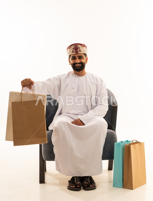 شراء المستلزمات و التبضع من المولات التجارية، الاثر الايجابي على النفسية، الاستفادة من العروض الموسمية و التخفيضات, بورتريه لرجل عربي خليجي عماني يرتدي الدشداشة و العمامة يجلس على الكرسى يمسك بيدية أكياس ورقية ملونة و ينظر الى الكاميرا، خلفية بيضاء