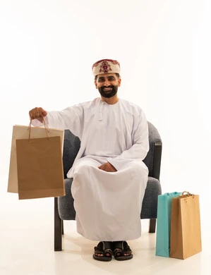شراء المستلزمات و التبضع من المولات التجارية، الاثر الايجابي على النفسية، الاستفادة من العروض الموسمية و التخفيضات, بورتريه لرجل عربي خليجي عماني يرتدي الدشداشة و العمامة يجلس على الكرسى يمسك بيدية أكياس ورقية ملونة و ينظر الى الكاميرا، خلفية بيضاء
