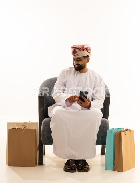 مفهوم التسوق الالكتروني، تصفح مواقع التواصل الاجتماعي، ايماءات السعادة و السرور , بورتريه لرجل عربي خليجي عماني يرتدي الدشداشة و العمامة يجلس على الكرسى يمسك الهاتف المحمول بيده و بجانبه أكياس ورقية , خلفية بيضاء