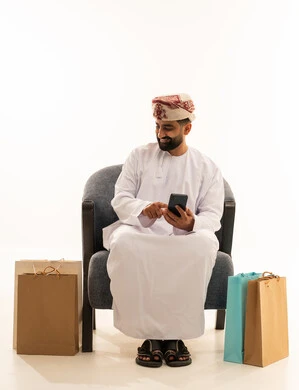 مفهوم التسوق الالكتروني، تصفح مواقع التواصل الاجتماعي، ايماءات السعادة و السرور , بورتريه لرجل عربي خليجي عماني يرتدي الدشداشة و العمامة يجلس على الكرسى يمسك الهاتف المحمول بيده و بجانبه أكياس ورقية , خلفية بيضاء