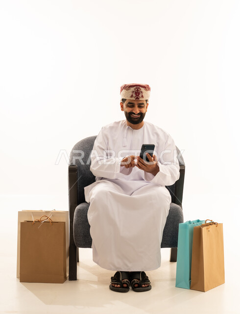 Browsing social media, gestures of happiness and joy, the concept of online shopping, a portrait of an Arab Gulf Omani man wearing a dishdasha and a turban sitting on a chair holding a mobile phone in his hand with paper bags beside him, white background.