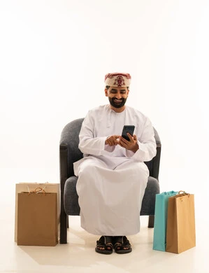 تصفح مواقع التواصل الاجتماعي، ايماءات السعادة و السرور، مفهوم التسوق الالكتروني , بورتريه لرجل عربي خليجي عماني يرتدي الدشداشة و العمامة يجلس على الكرسى يمسك الهاتف المحمول بيده و بجانبه أكياس ورقية , خلفية بيضاء