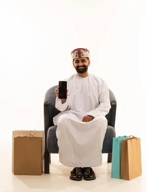 عرض شاشة سوداء فارغة عبر الجوال، شراء المنتجات و الأغراض عبر الانترنت , بورتريه لرجل عربي خليجي عماني يرتدي الدشداشة و العمامة يجلس على الكرسى يمسك الهاتف المحمول بيده و بجانبه أكياس ورقية ، خلفية بيضاء