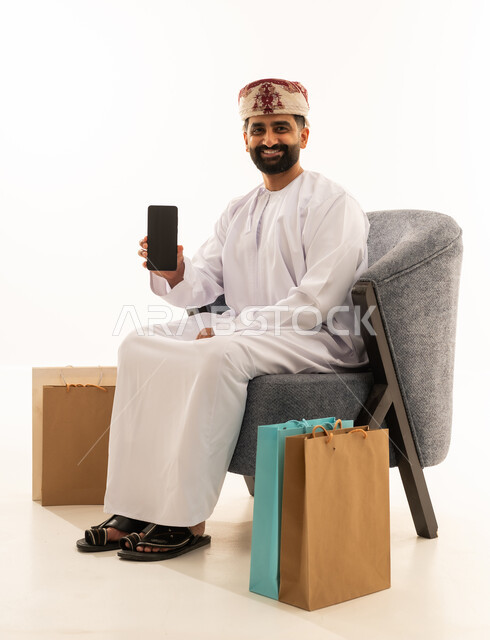 شراء المنتجات و الأغراض عبر الانترنت , بورتريه لرجل عربي خليجي عماني يرتدي الدشداشة و العمامة يجلس على الكرسى يمسك الهاتف المحمول بيده و بجانبه أكياس ورقية , عرض شاشة سوداء فارغة عبر الجوال ، خلفية بيضاء