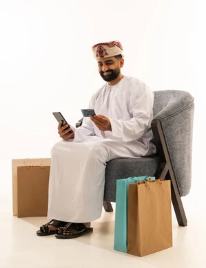 مفهوم الدفع والتسديد عن بعد عبر تطبيقات الجوال، التسوق الالكتروني عبر الانترنت، استخدام البطاقة البنكية الالكترونية، بورتريه لرجل عربي خليجي عماني يرتدي الدشداشة و العمامة يجلس على الكرسى بجانب حقائب التسوق يحمل الهاتف المحمول، خلفية بيضاء