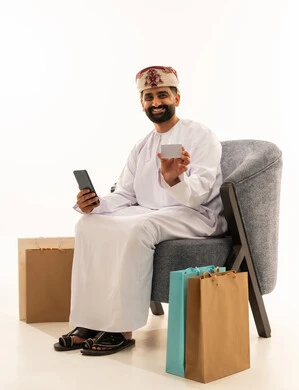 التسوق الالكتروني عبر الانترنت، مفهوم الدفع والتسديد عن بعد عبر تطبيقات الجوال، استخدام البطاقة البنكية الالكترونية، بورتريه لرجل عربي خليجي عماني يرتدي الدشداشة و العمامة يجلس على الكرسى بجانب حقائب التسوق يحمل الهاتف المحمول، خلفية بيضاء
