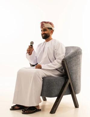 Delivering speeches and words using a loudspeaker, engaging in dialogues and discussions, a portrait of an Arab Gulf Omani man wearing a dishdasha and a turban sitting on a chair holding a loudspeaker in his hand, white background.