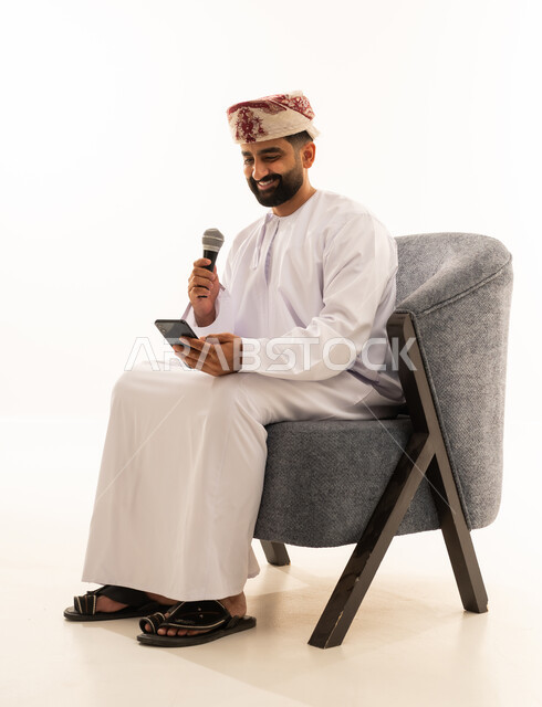 Practicing the hobby of singing for fun and entertainment, artistic talents in the world of music, musical theater art, presenting the theatrical performance, a portrait of an Arab Gulf Omani man wearing a dishdasha and a turban sitting on a chair holding a microphone and a mobile phone, white background.