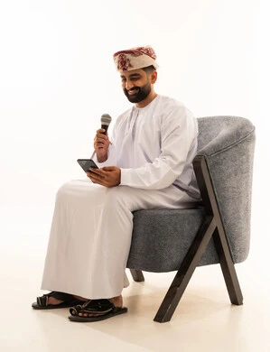 Practicing the hobby of singing for fun and entertainment, artistic talents in the world of music, musical theater art, presenting the theatrical performance, a portrait of an Arab Gulf Omani man wearing a dishdasha and a turban sitting on a chair holding a microphone and a mobile phone, white background.