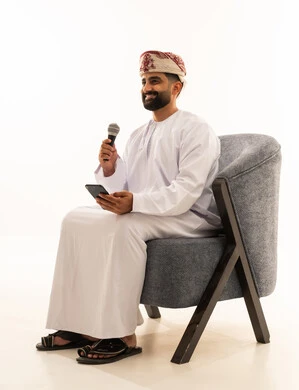 Artistic talents in the world of music, practicing the hobby of singing for fun and entertainment, musical theater art, presenting the theatrical performance, a portrait of an Arab Gulf Omani man wearing a dishdasha and a turban sitting on a chair holding a microphone and a mobile phone, white background.