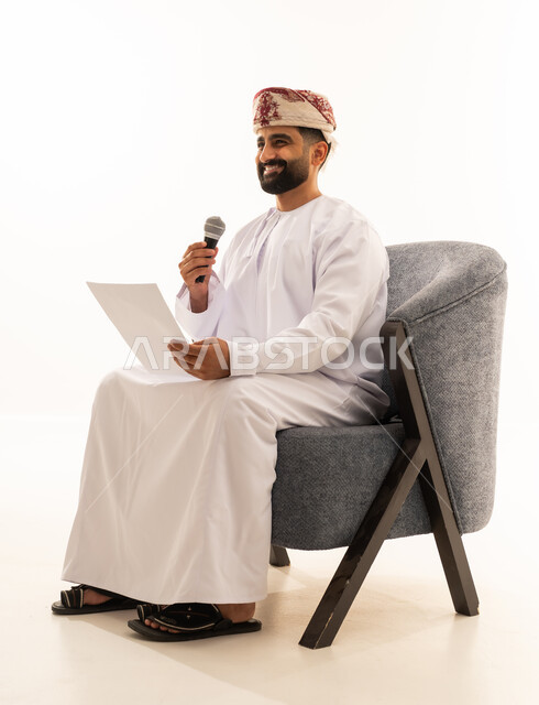 اعداد التقارير الخاصة بالعمل، تقديم الفقرات باستخدام الميكروفون، متابعة ومراجعة الخطط والمشاريع المكتبية، بورتريه لرجل عربي خليجي عماني يرتدي الدشداشة و العمامة يجلس على الكرسى ينظر الى الاوراق البيضاء يمسك بيده المايك, التدقيق في قراءة الملاحظات، خلفية بيضاء