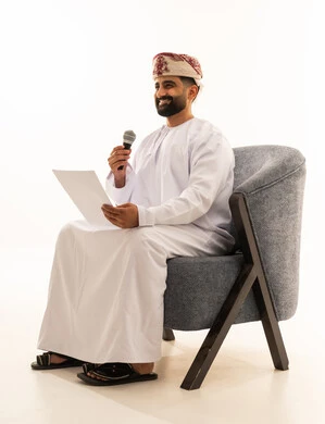 اعداد التقارير الخاصة بالعمل، تقديم الفقرات باستخدام الميكروفون، متابعة ومراجعة الخطط والمشاريع المكتبية، بورتريه لرجل عربي خليجي عماني يرتدي الدشداشة و العمامة يجلس على الكرسى ينظر الى الاوراق البيضاء يمسك بيده المايك, التدقيق في قراءة الملاحظات، خلفية بيضاء