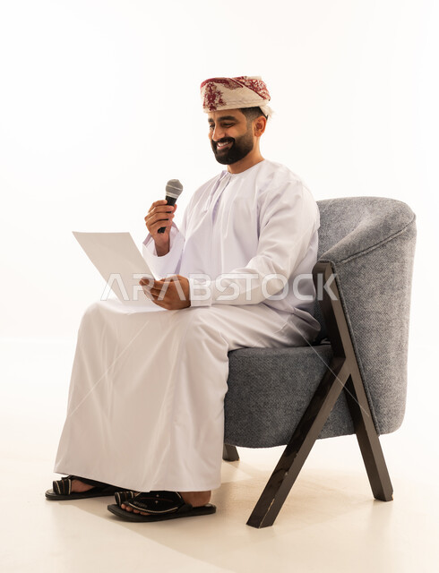 Presenting segments using the microphone, preparing work-related reports, following up and reviewing office plans and projects, a portrait of an Arab Gulf Omani man wearing a dishdasha and a turban sitting on a chair looking at white papers holding a microphone in his hand, proofreading notes, white background.