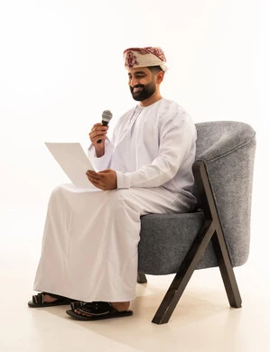 Presenting segments using the microphone, preparing work-related reports, following up and reviewing office plans and projects, a portrait of an Arab Gulf Omani man wearing a dishdasha and a turban sitting on a chair looking at white papers holding a microphone in his hand, proofreading notes, white background.