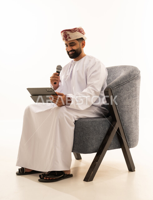 مواهب غنائية فنية، تسجيل مقطع صوتي غنائي، الاندماج مع الأغنية، بورتريه لرجل عربي خليجي عماني يرتدي الدشداشة و العمامة يجلس على الكرسى يقوم بالغناء باستخدام الميكروفون يمسك بيدية التابلت بتعابير الحماس، خلفية بيضاء