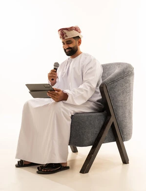مواهب غنائية فنية، تسجيل مقطع صوتي غنائي، الاندماج مع الأغنية، بورتريه لرجل عربي خليجي عماني يرتدي الدشداشة و العمامة يجلس على الكرسى يقوم بالغناء باستخدام الميكروفون يمسك بيدية التابلت بتعابير الحماس، خلفية بيضاء