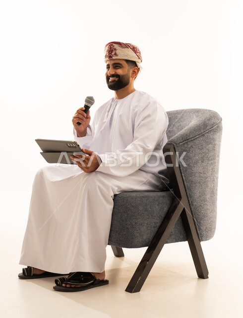 تسجيل مقطع صوتي غنائي، مواهب غنائية فنية، الاندماج مع الأغنية، بورتريه لرجل عربي خليجي عماني يرتدي الدشداشة و العمامة يجلس على الكرسى يقوم بالغناء باستخدام الميكروفون يمسك بيدية التابلت بتعابير الحماس، خلفية بيضاء