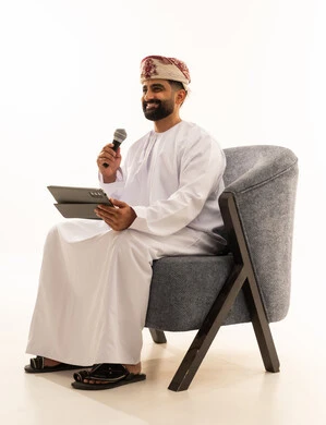 تسجيل مقطع صوتي غنائي، مواهب غنائية فنية، الاندماج مع الأغنية، بورتريه لرجل عربي خليجي عماني يرتدي الدشداشة و العمامة يجلس على الكرسى يقوم بالغناء باستخدام الميكروفون يمسك بيدية التابلت بتعابير الحماس، خلفية بيضاء