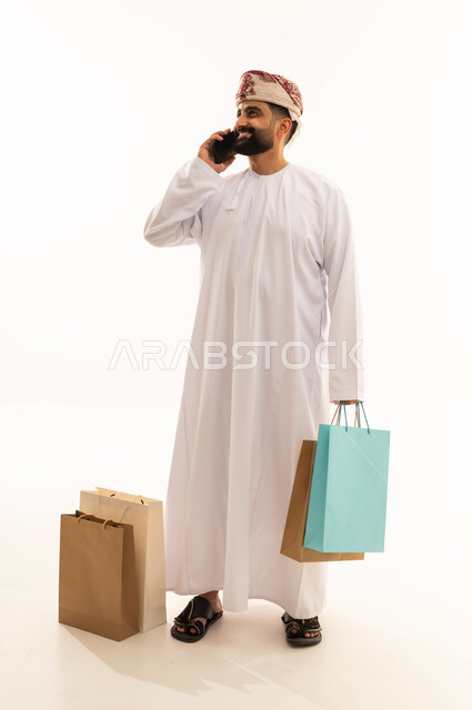 اجراء مكالمة هاتفية عبر الجوال، مفهوم التسوق عن بعد, عمليات الشراء الالكتروني عبر الانترنت,بورتريه لرجل عربي خليجي عماني يرتدي الدشداشة و العمامة يمسك بيدية أكياس ورقية ملونة، التبضع والاستفادة من العروض والتخفيضات, الجسم بالطول الكامل, خلفية بيضاء