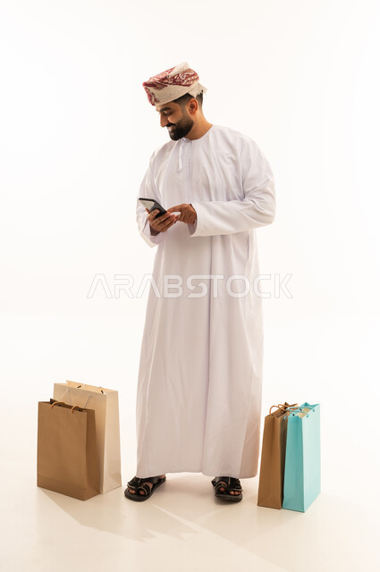 Browsing social media, the concept of online shopping, a portrait of an Arab Gulf Omani man wearing a dishdasha and a turban holding a mobile phone in his hand with paper bags beside him, full body, white background.