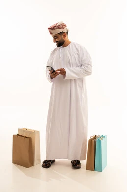 تصفح مواقع التواصل الاجتماعي ، مفهوم التسوق الالكتروني, بورتريه لرجل عربي خليجي عماني يرتدي الدشداشة و العمامة يمسك الهاتف المحمول بيده و بجانبه أكياس ورقية, الجسم بالطول الكامل، خلفية بيضاء