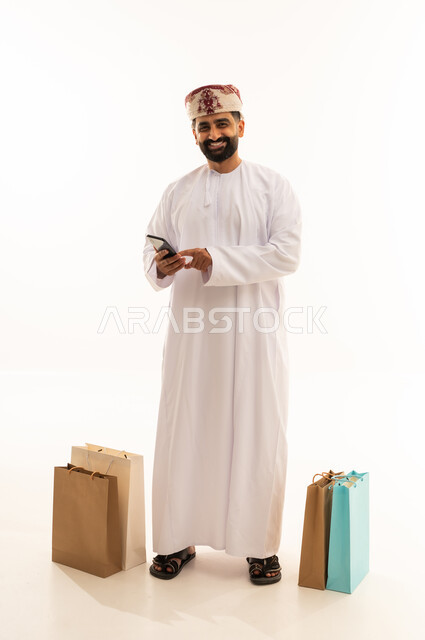 The concept of electronic shopping, browsing social media sites, a portrait of an Arab Gulf Omani man wearing a dishdasha and turban holding a mobile phone in his hand with paper bags beside him, full body, white background.
