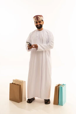 مفهوم التسوق الالكتروني ,تصفح مواقع التواصل الاجتماعي , بورتريه لرجل عربي خليجي عماني يرتدي الدشداشة و العمامة يمسك الهاتف المحمول بيده و بجانبه أكياس ورقية, الجسم بالطول الكامل، خلفية بيضاء