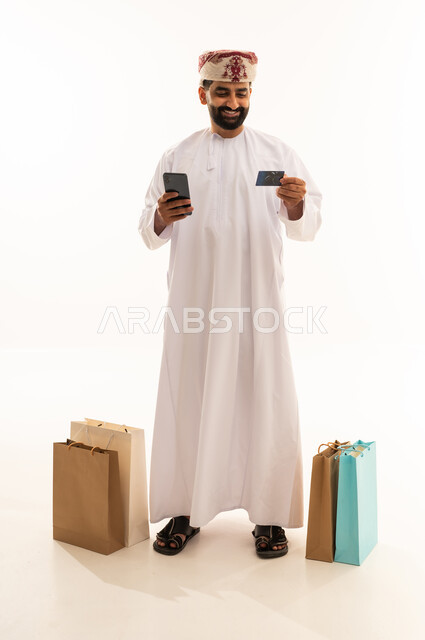 مفهوم التسوق عن بعد، الشراء والدفع عن طريق بطاقة الإئتمان البنكية، إتمام عملية الطلبات باستخدام الهاتف المحمول, بورتريه لرجل عربي خليجي عماني يرتدي الدشداشة و العمامة يقف بجانب حقائب ورقية يمسك بيده الجوال و كارت بنكي , التبضع والاستفادة من العروض والتخفيضات, الجسم بالطول الكامل، خلفية بيضاء