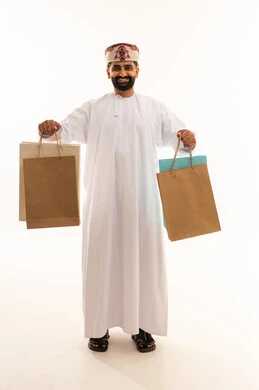 الاُثر الايجابي للتسوق، شراء الهدايا والمستلزمات، الاستفادة من العروض والتخفيضات الموسميه، بورتريه لرجل عربي خليجي عماني يرتدي الدشداشة و العمامة يحمل بيده أكياس ورقية ملونة بإيماءات السرور والسعادة، الجسم بالطول الكامل، خلفية بيضاء