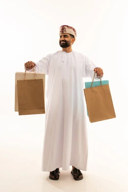Buying gifts and supplies, benefiting from seasonal offers and discounts, the positive impact of shopping, a portrait of an Arab Gulf Omani man wearing a dishdasha and a turban holding colorful paper bags in gestures of joy and happiness, full-body, white background.