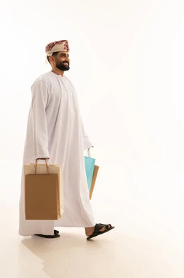 Taking advantage of offers and discounts, the joy of shopping and acquiring necessities and gifts, a side portrait of an Arab Gulf Omani man wearing a dishdasha and a turban holding colorful paper bags in his hand with gestures of joy and happiness, full-body, white background.