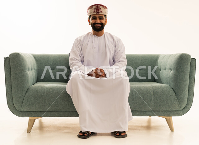 ايماءات وجه تدل على السعادة و السرور,، مفهوم الرجولة والثقة بالنفس، الاهتمام بالأناقة والمظهر الخارجي، بورتريه لرجل عربي خليجي عماني يرتدي الدشداشة و العمامة يجلس على الاريكة، خلفية بيضاء