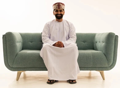 ايماءات وجه تدل على السعادة و السرور,، مفهوم الرجولة والثقة بالنفس، الاهتمام بالأناقة والمظهر الخارجي، بورتريه لرجل عربي خليجي عماني يرتدي الدشداشة و العمامة يجلس على الاريكة، خلفية بيضاء