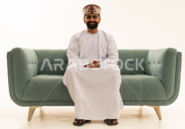 النظر للكاميرا بتعابير السرور، مفهوم الرجولة والثقة بالنفس، الاهتمام بالأناقة والمظهر الخارجي، بورتريه لرجل عربي خليجي عماني يرتدي الدشداشة و العمامة يجلس على الاريكة، خلفية بيضاء