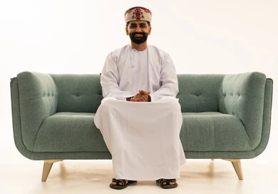 Looking at the camera with expressions of joy, the concept of masculinity and self-confidence, attention to elegance and appearance, a portrait of an Arab Gulf Omani man wearing a dishdasha and a turban sitting on a sofa, white background.