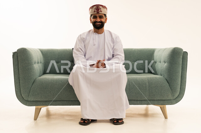 مفهوم الرجولة والثقة بالنفس، الاهتمام بالأناقة والمظهر الخارجي، بورتريه لرجل عربي خليجي عماني يرتدي الدشداشة و العمامة يجلس على الاريكة ينظر الى الكاميرا بتعابير السرور، خلفية بيضاء